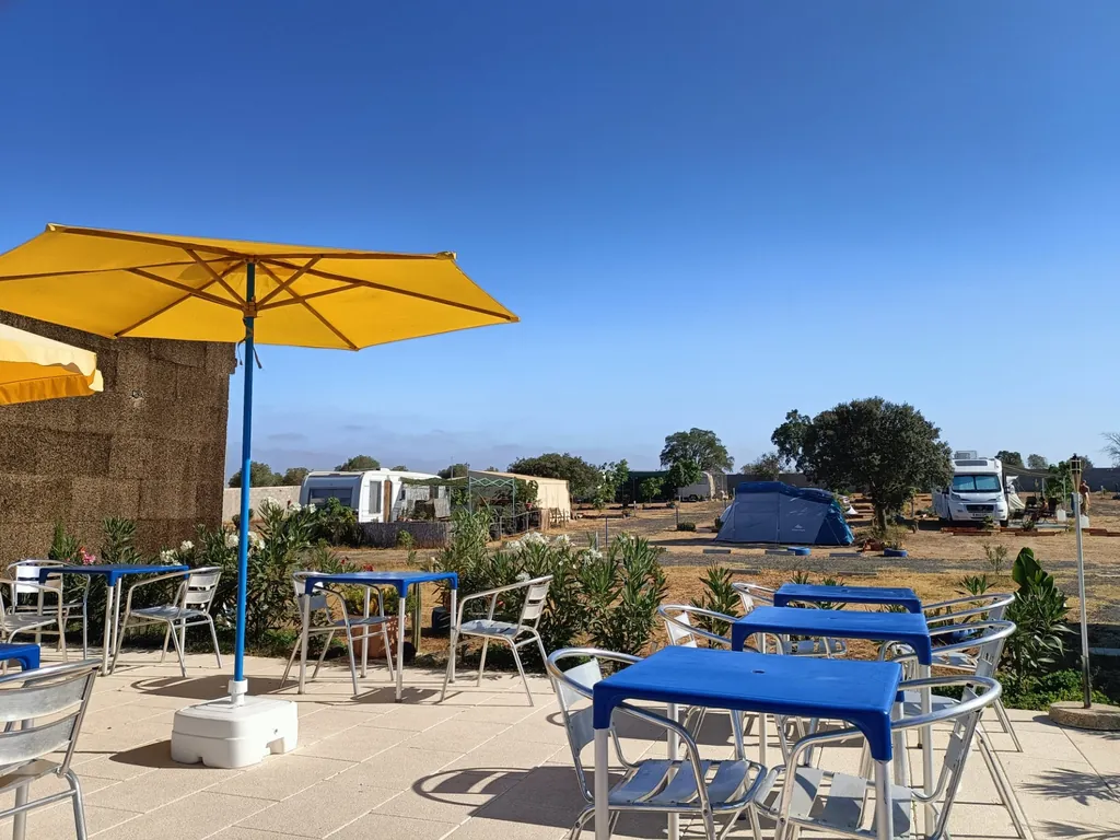 Área de bar con mesas, sombrilla amarilla y vistas a parcelas del Camping Terra Nua - Parque de Campismo e Caravanismo Naturista, Aljustrel (Beja)
