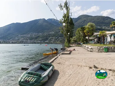 Playa junto al mar con bungalows, montaña al fondo del Camping Rivabella, Tenero (Tesino)