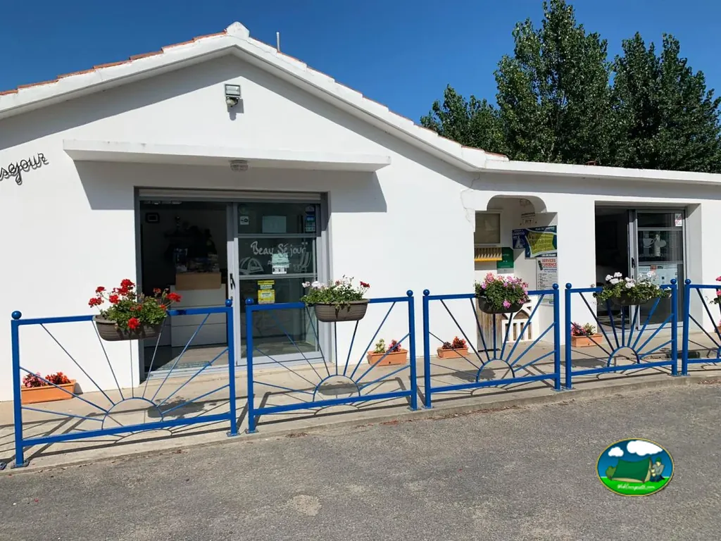 recepción del camping Beau Séjour con fachada blanca, valla azul y jardineras de flores