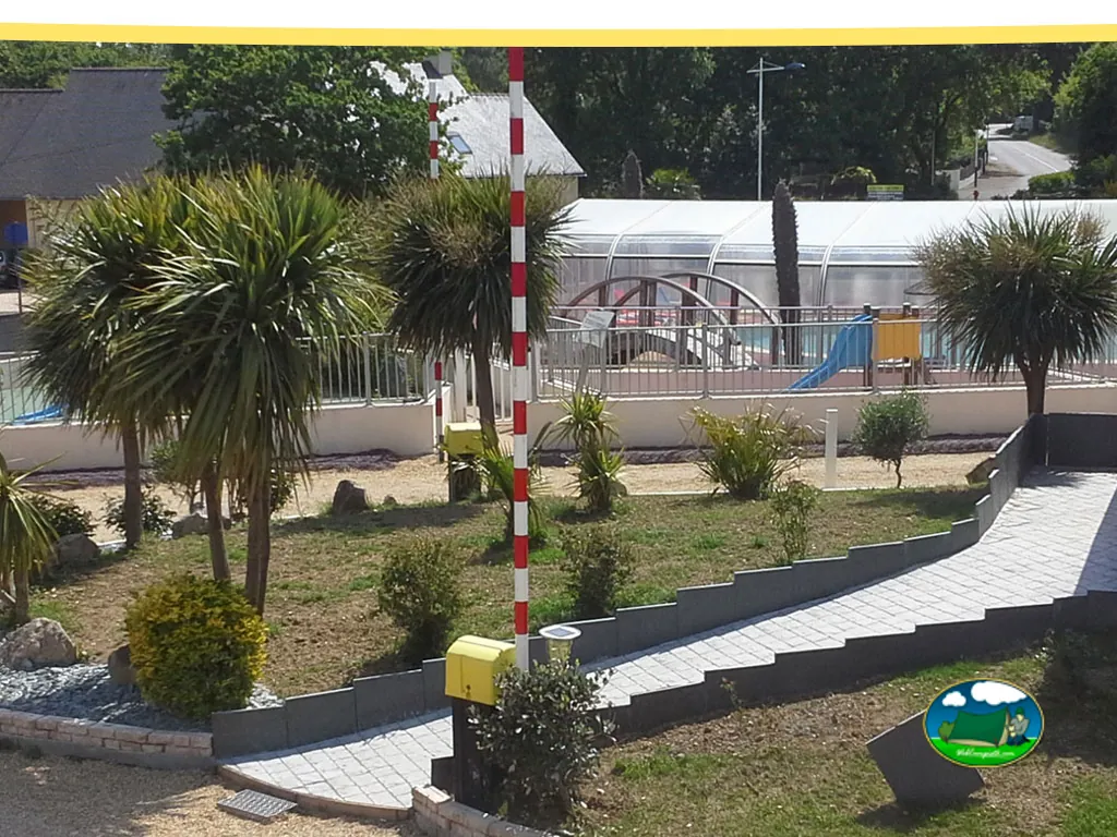 Jardín del camping con palmeras, piscina cubierta con tobogán y zona infantil al fondo.