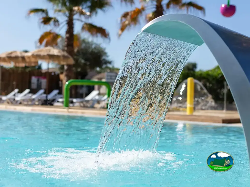 Primer plano de una cascada artificial en la piscina, con palmeras y hamacas de fondo.