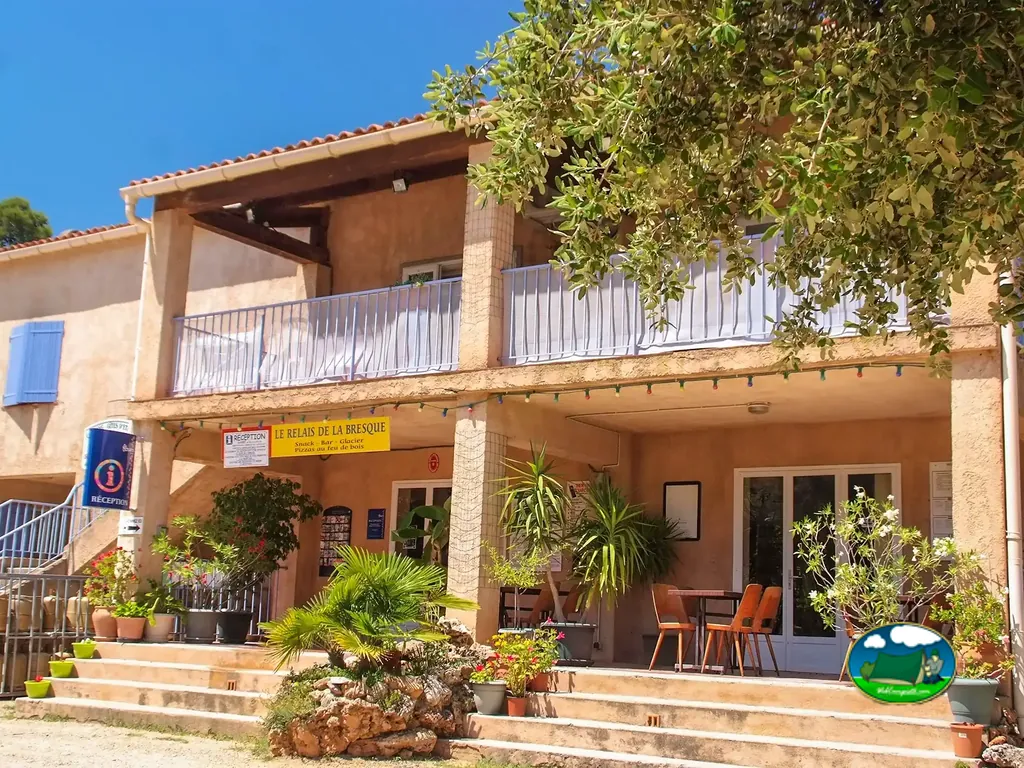 Fachada de la recepción y el snack-bar del camping, con terraza, plantas y mesas al sol.