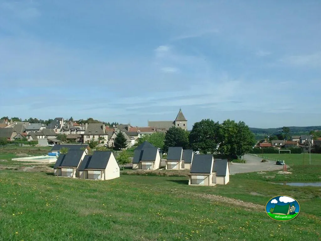 hileras de pequeños chalets de tejado oscuro junto a la piscina, con el pueblo y la iglesia de Marcolès al fondo