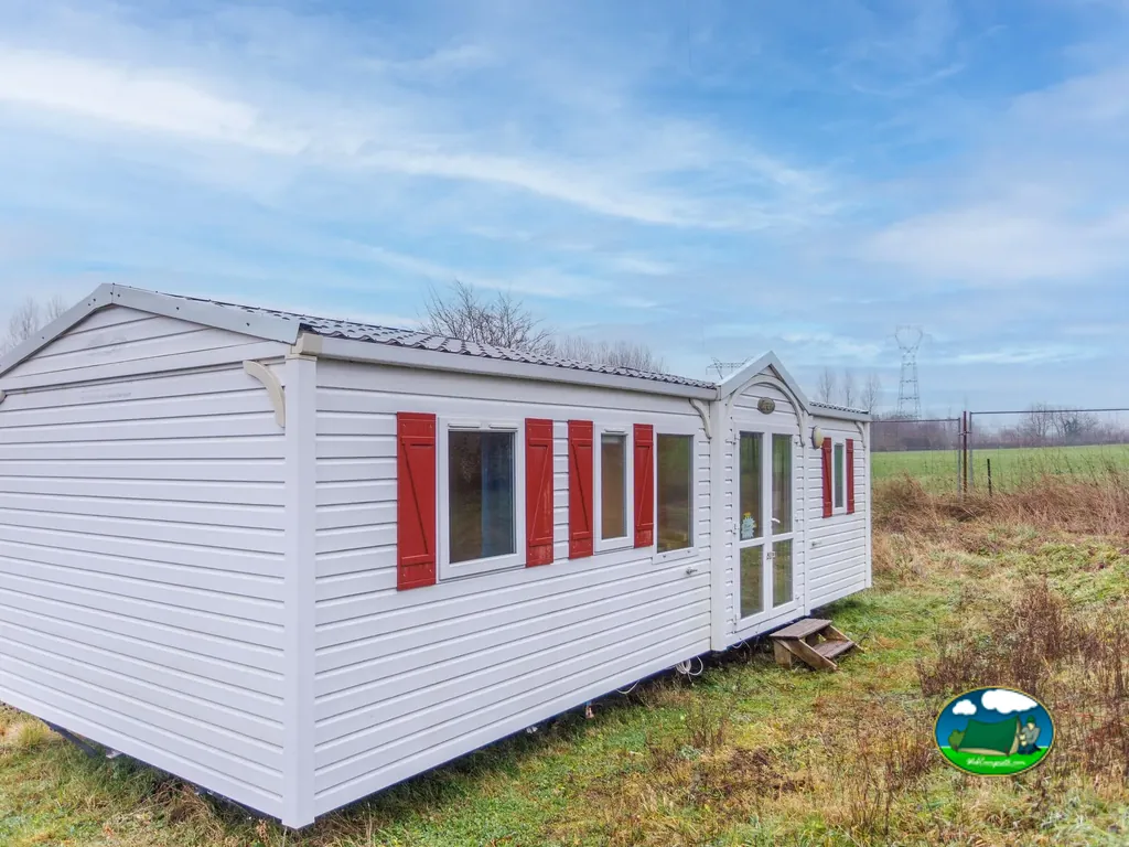 mobil-home blanco con contraventanas rojas en una parcela de hierba junto a un campo en Gommegnies