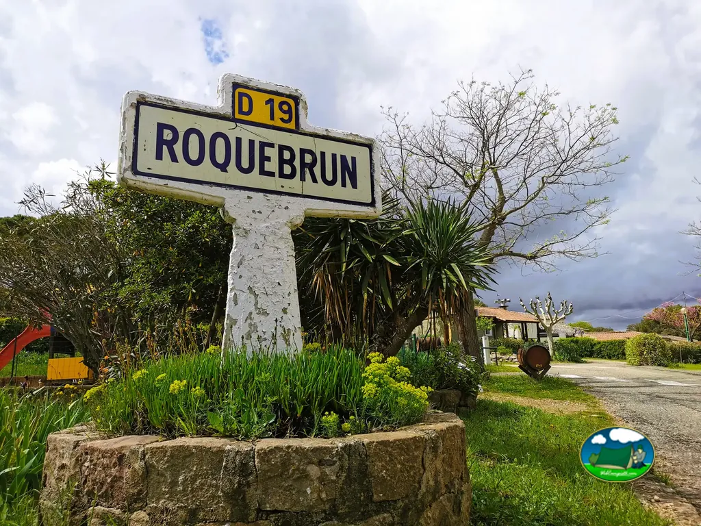 cartel de carretera de Roquebrun junto a un tobogán infantil y edificios del camping bajo cielo nuboso