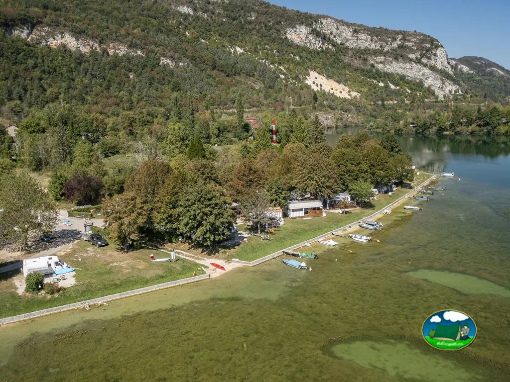 vista aérea del camping junto a un lago con parcelas arboladas, embarcaciones y montañas al fondo en Condes