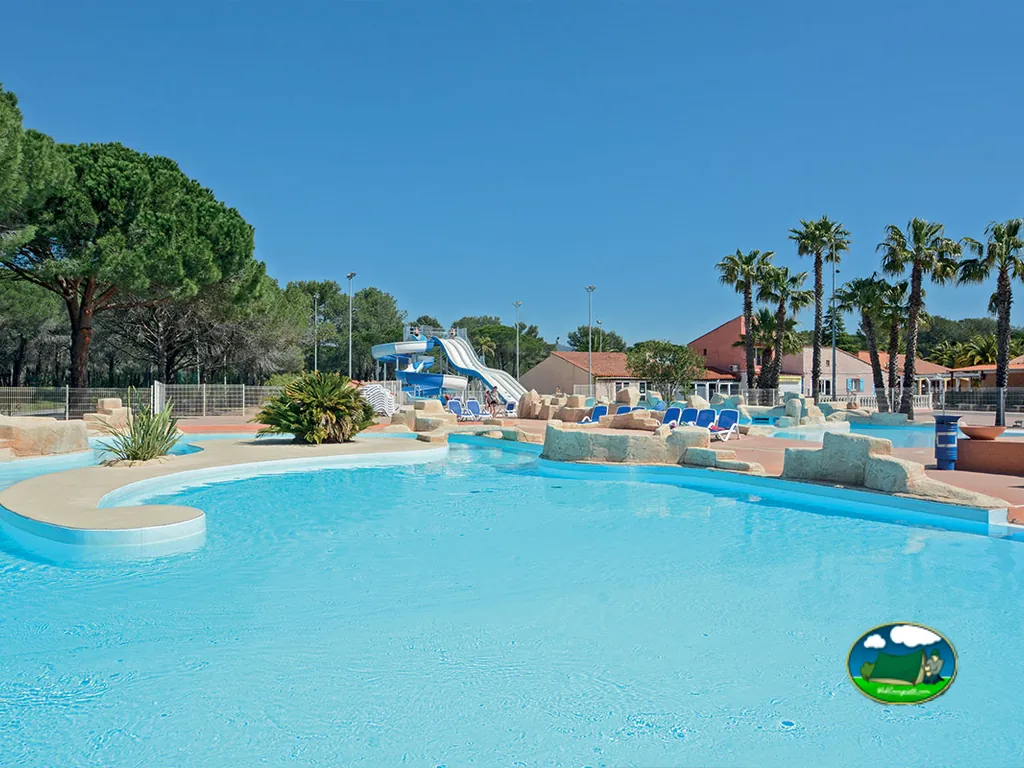 piscina con tobogán de agua, tumbonas azules y palmeras junto a los edificios del camping