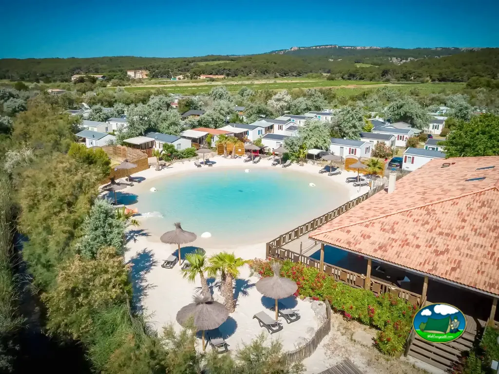 vista aérea de una piscina lago con arena blanca, palmeras y sombrillas de paja rodeada de bungalows en Narbonne