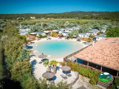 vista aérea de una piscina lago con arena blanca, palmeras y sombrillas de paja rodeada de bungalows en Narbonne