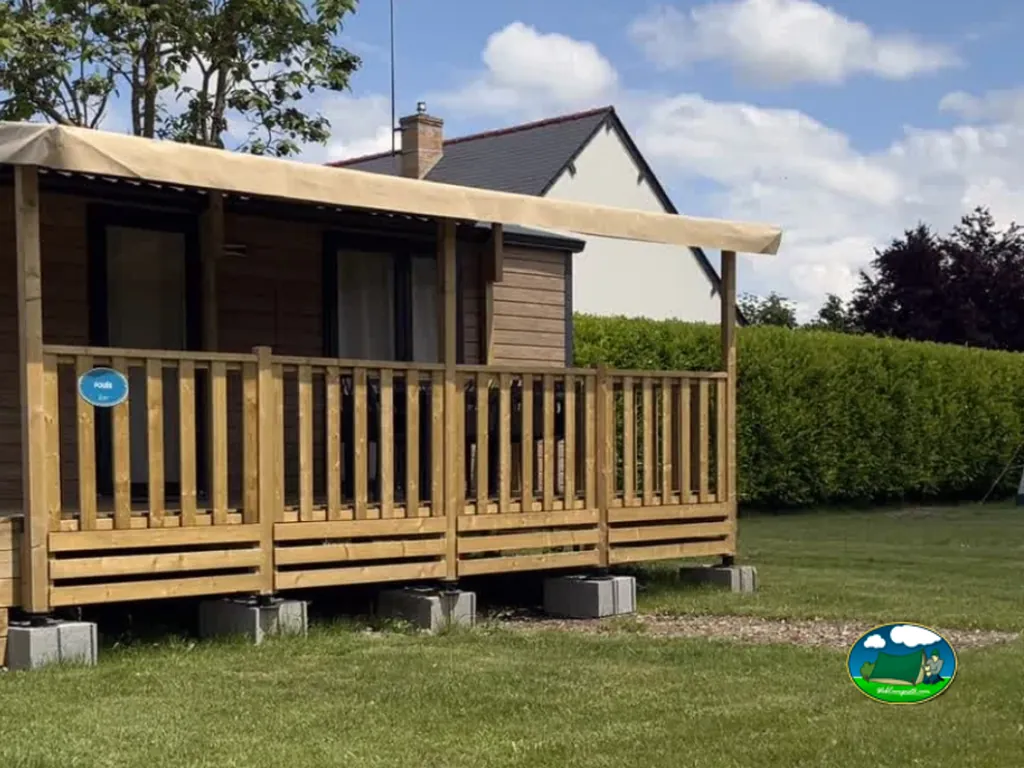 chalet de madera con terraza cubierta y toldo en el camping, casa vecina al fondo bajo cielo despejado
