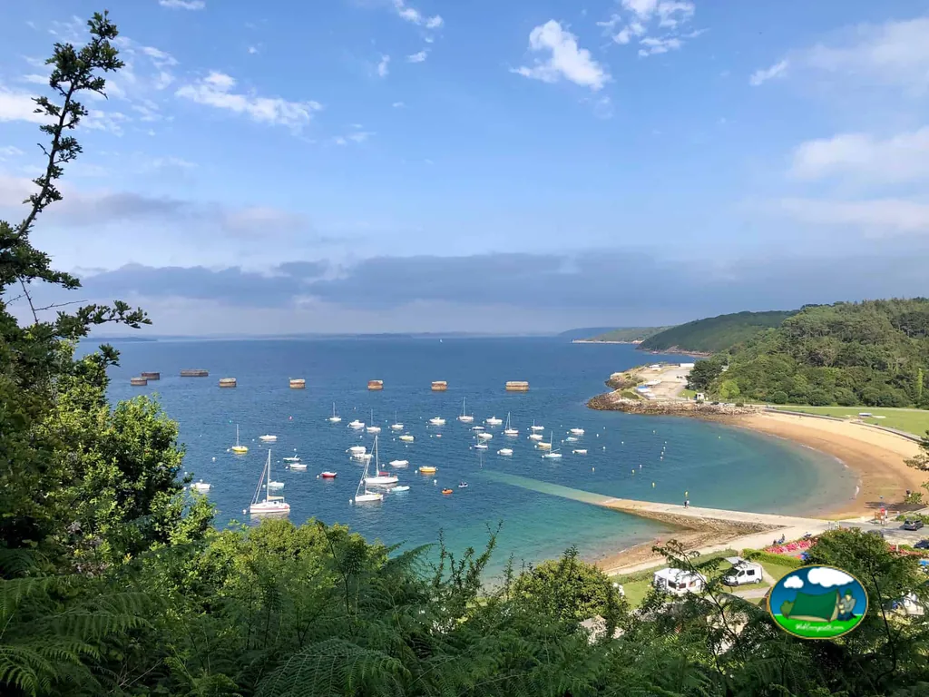 vista de la bahía con veleros fondeados y playa, con autocaravanas del camping asomando entre la vegetación en primer plano cerca de Lanvéoc
