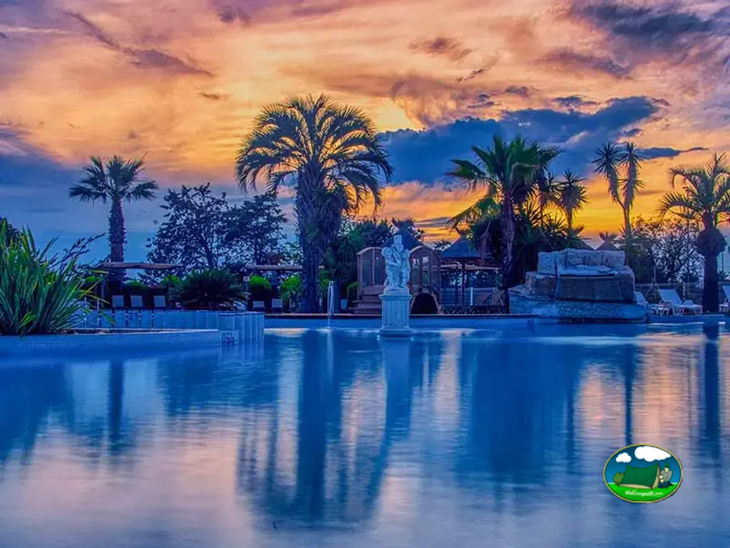 piscina del camping al atardecer con palmeras, estatua y cielo anaranjado reflejado en el agua