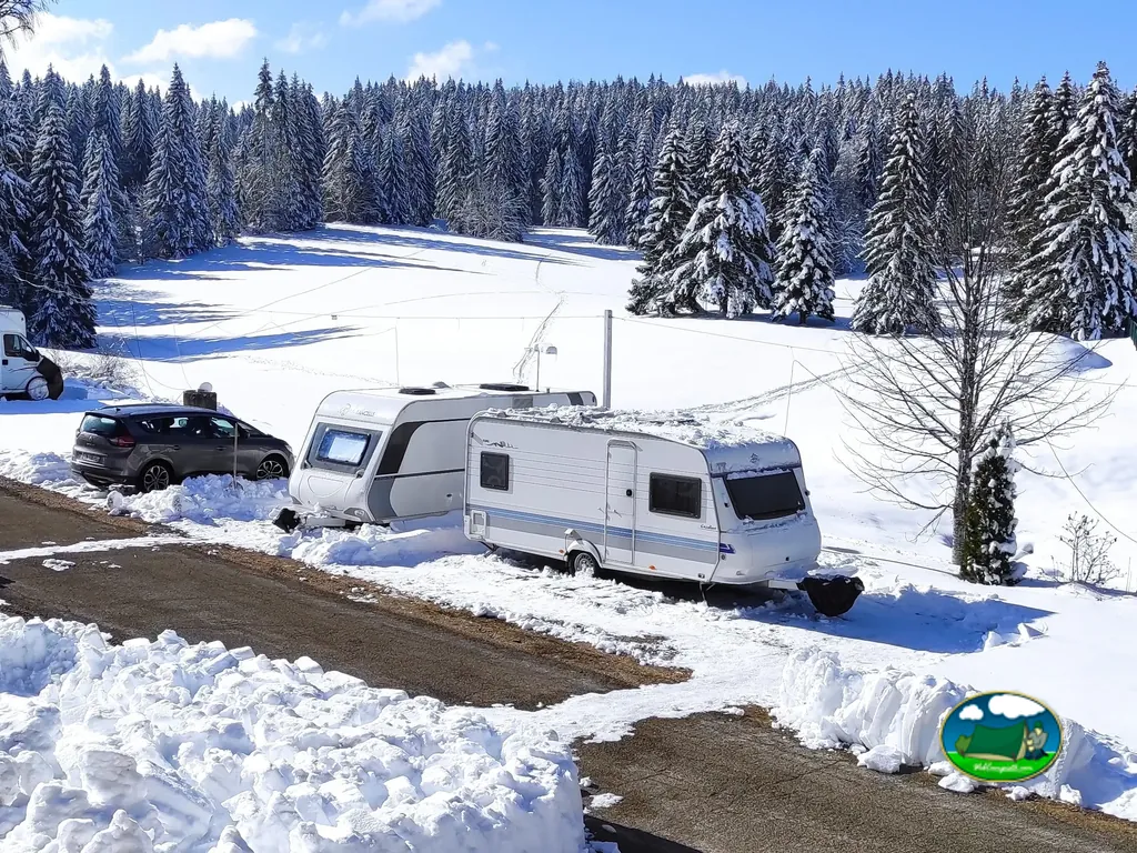 caravanas nevadas aparcadas junto a un coche en la parcela del camping rodeada de abetos nevados en Prémanon