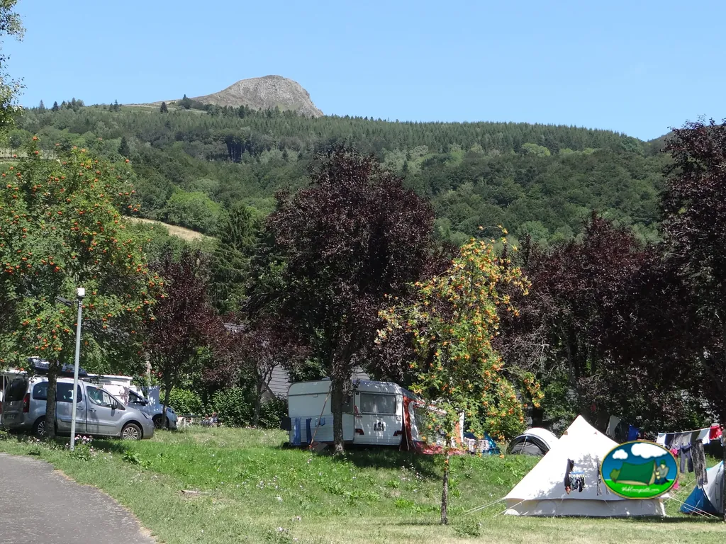 parcelas del camping con caravana y tienda de campaña bajo árboles con un puy volcánico al fondo en La Bourboule