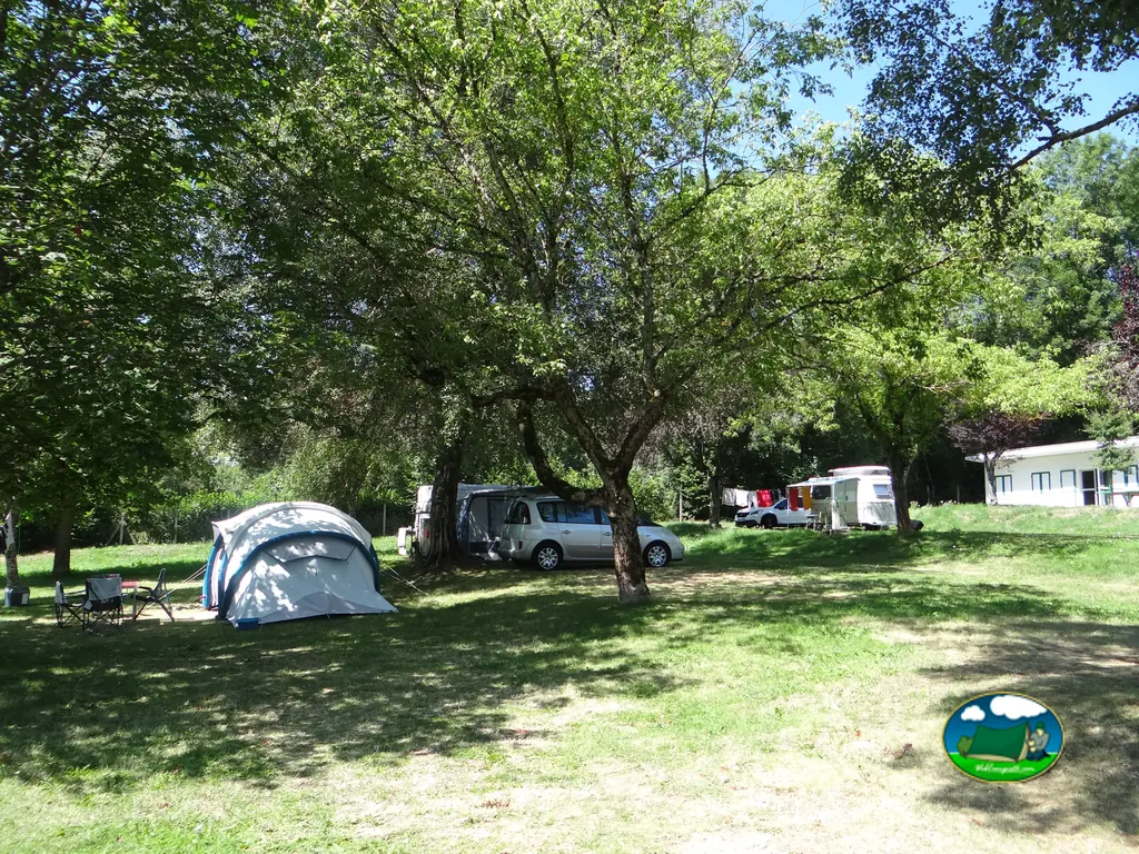 parcela sombreada del camping con tienda, turismo y caravana bajo grandes árboles junto al edificio de recepción