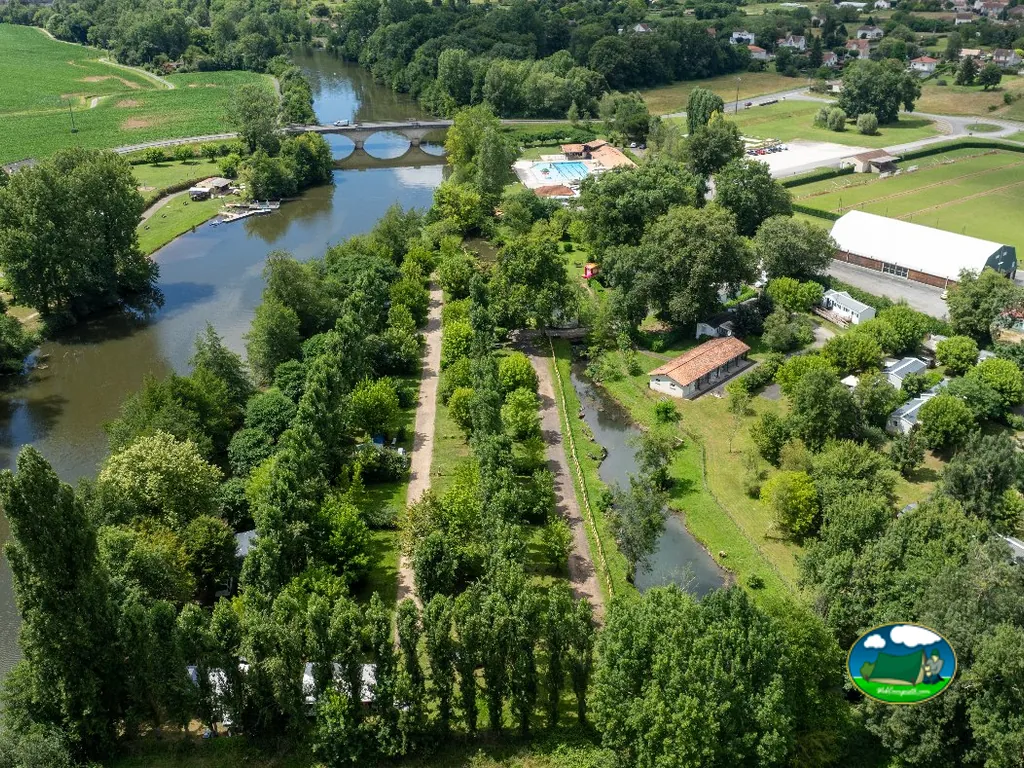 vista aérea del camping junto al río con piscina, puente de piedra y parcelas arboladas en Neuvic