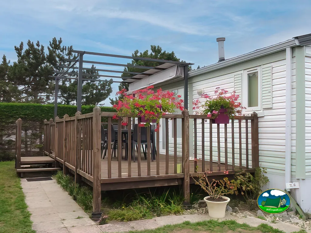 terraza de un mobil-home con pérgola, macetas de flores rojas y mesa exterior