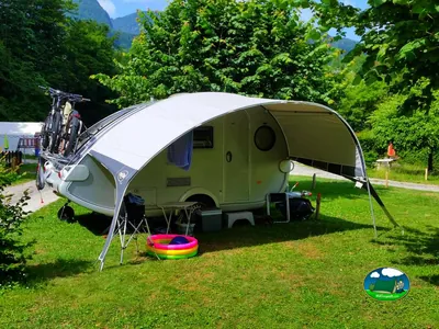 caravana con toldo desplegado, bicicletas y una piscina hinchable infantil sobre césped con montañas al fondo