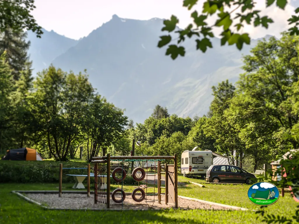 zona de juegos infantiles junto a una caravana y un coche aparcados entre árboles con montañas al fondo