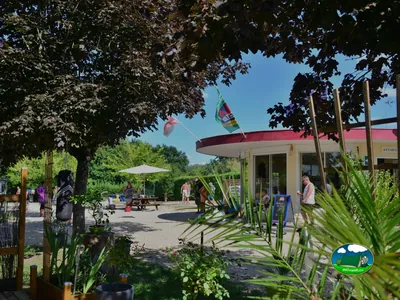 edificio de recepción con banderas junto a una terraza con mesas y varias personas alrededor