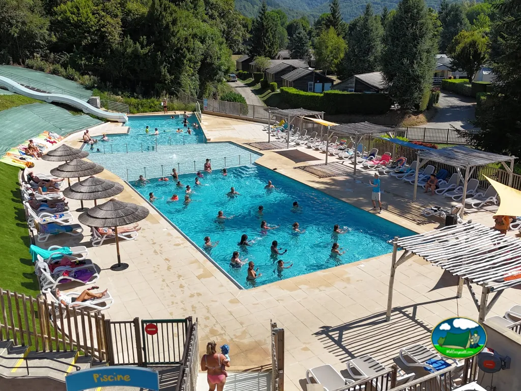 piscina concurrida con tobogán y bañistas vista desde arriba, chalets entre montañas boscosas en Saint-Geniez-d'Olt