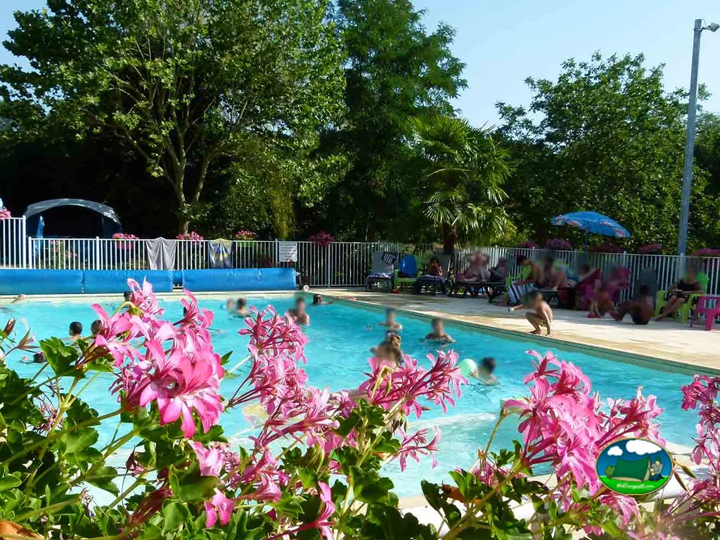 piscina con bañistas tras un macizo de flores rosas y palmeras en el camping Le Pigeonnier