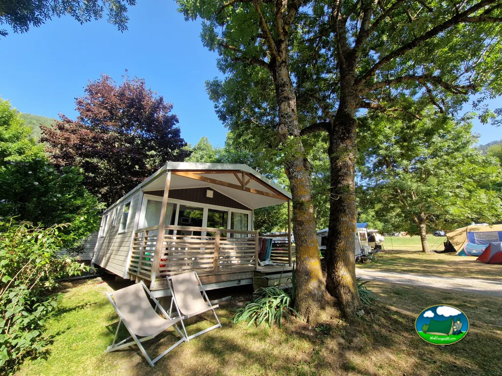 bungaló de madera bajo grandes árboles junto a caravanas y tiendas en el camping Le Ruisseau