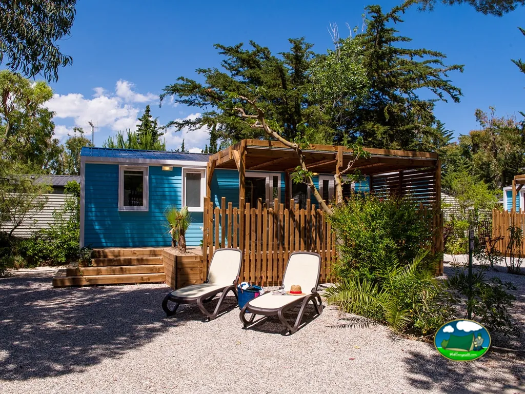 mobil-home azul con terraza de madera, pérgola y tumbonas en un jardín arbolado en Le Barcarès