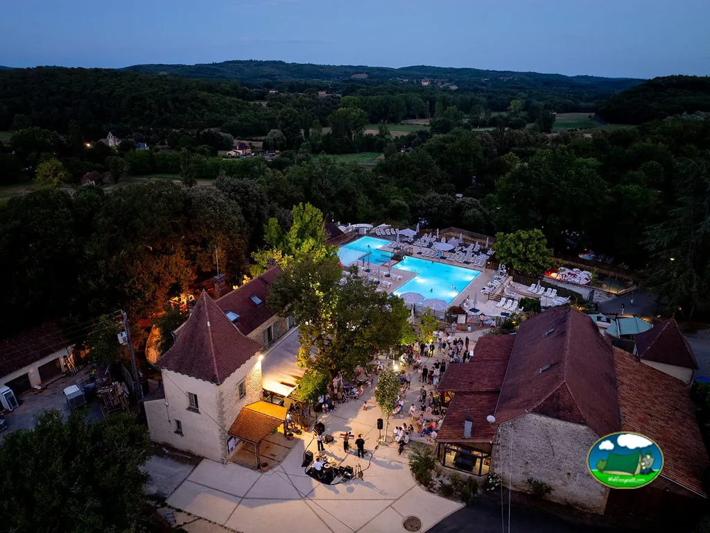 vista aérea nocturna del camping con piscina iluminada, edificio de piedra tipo castillo y público reunido para una actuación musical