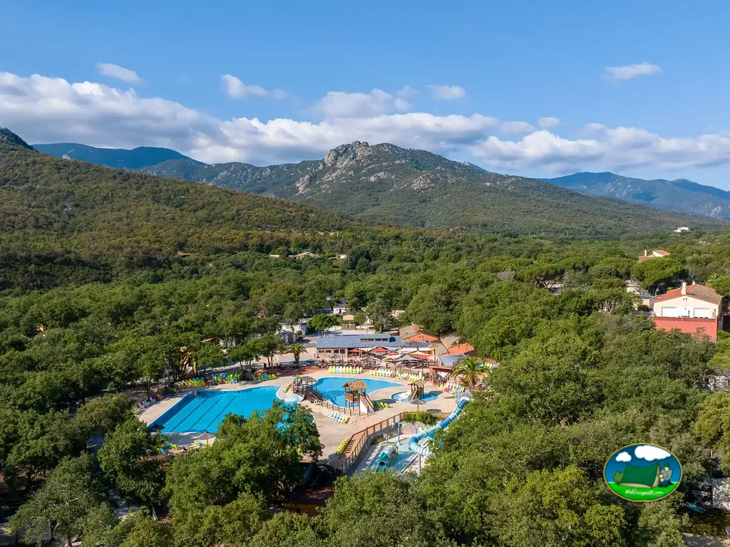 vista aérea diurna del parque acuático del camping con toboganes, piscinas y restaurante entre árboles y montañas al fondo