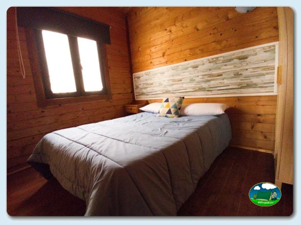 Cabaña de madera con cama doble y ventana del Camping Somo Parque, Ribamontán al Mar (Cantabria)