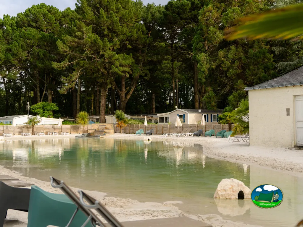 piscina de fondo arenoso con tumbonas rodeada de casas móviles y pinos