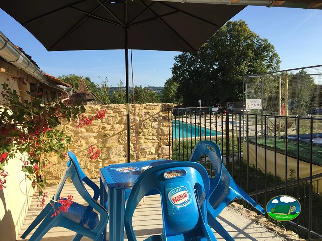 terraza con sillas de plástico bajo sombrilla junto a la piscina y la pista deportiva vistas entre la valla