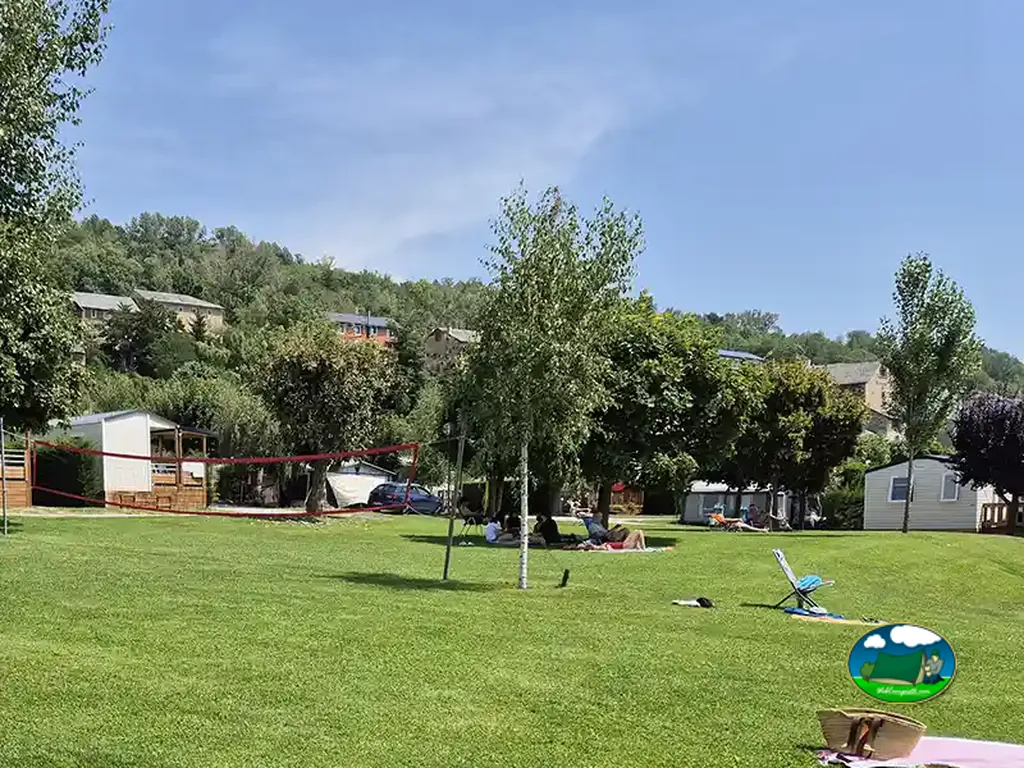 césped del camping con red de voleibol, personas descansando bajo un árbol y bungalós al fondo en Enveitg