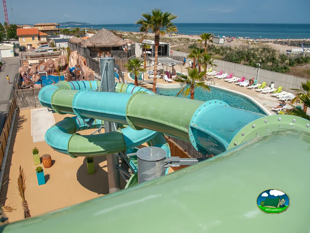 vista elevada de tobogán acuático tubular junto a piscina con tumbonas de colores y el mar al fondo