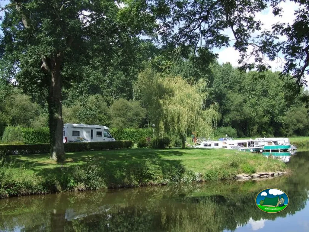 parcela de césped con autocaravana bajo sauce junto al canal con embarcaciones amarradas