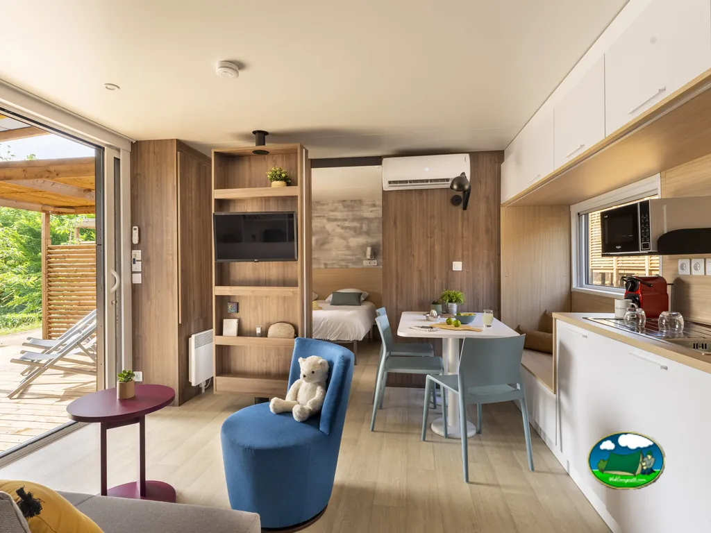 salón y cocina de un mobil-home con sofá, mesa y un osito de peluche sobre un sillón azul