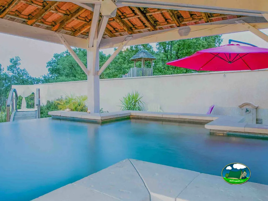 piscina cubierta por un porche de tejas con sombrilla roja y parque infantil al fondo en el camping Domaine des Chênes en Les Vans