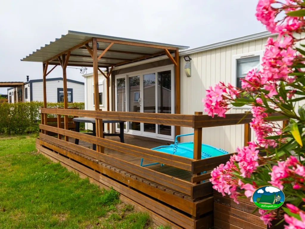 terraza de madera de un mobile home con mesa, piscina portátil y flores rosas en Le Boulou