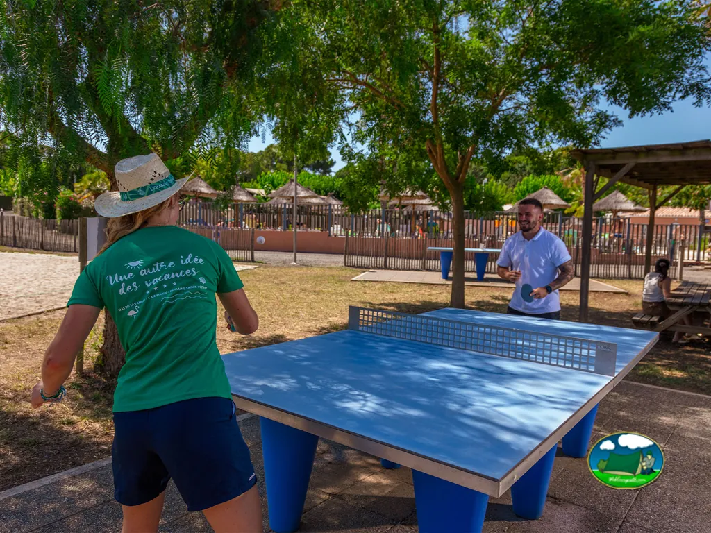 dos personas jugando al ping pong bajo los árboles cerca de la piscina del camping