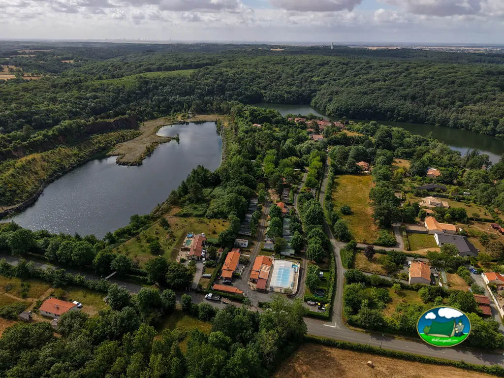vista aérea de un lago rodeado de bosque con el camping y su piscina al fondo en MERVENT
