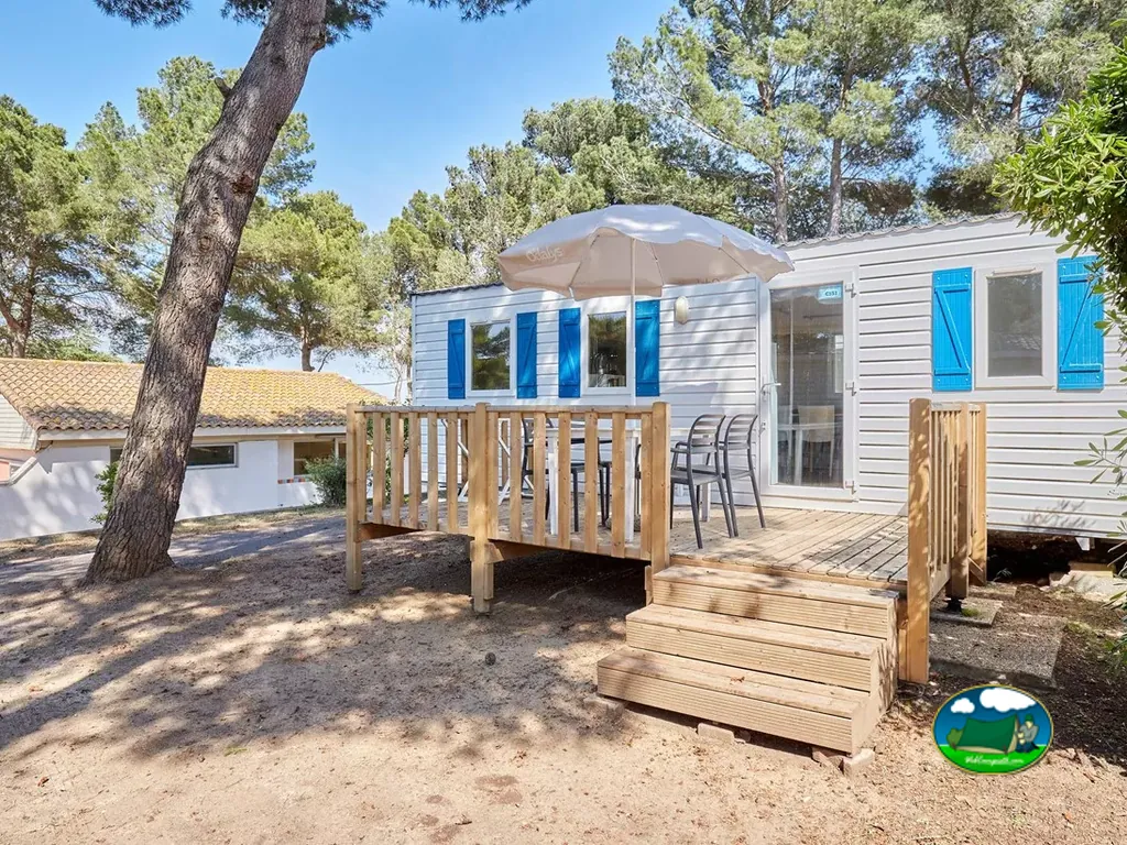 mobil-home con terraza de madera, mesa y sombrilla bajo pinos en Agde