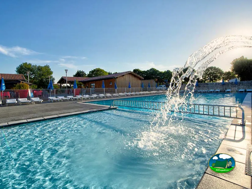 piscina con chorro de agua en primer plano y edificio de madera al fondo en el camping de La Flotte