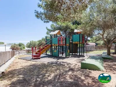 Parque infantil con tobogán y estructuras de juego bajo árboles del Camping Son Bou, Alaior (Menorca)